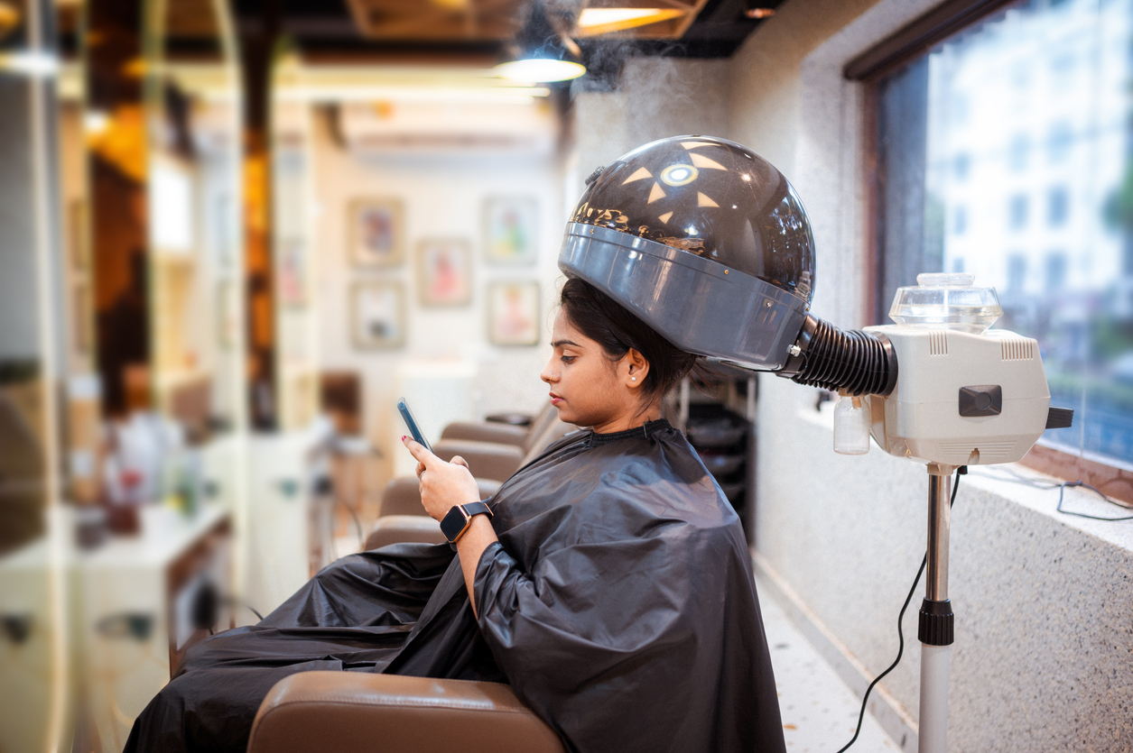 Client using a smartphone while getting hair treatment, highlighting the convenience of mobile apps and online booking platforms for salons.