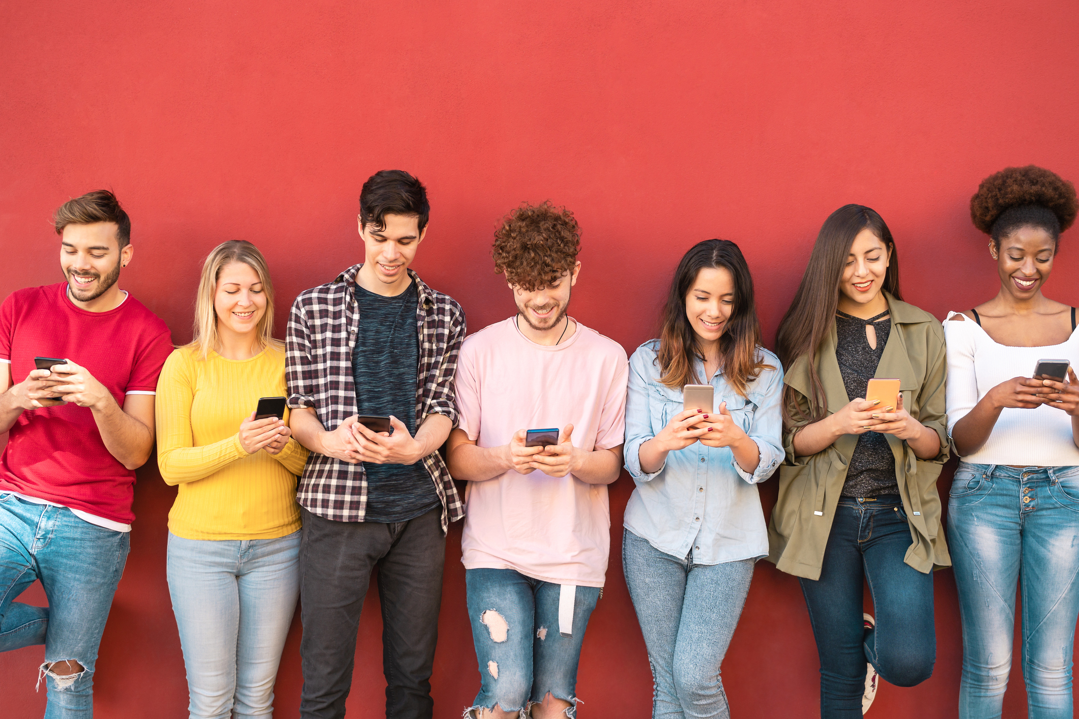 A diverse group of young adults standing against a red wall, all smiling while using their smartphones to send or read SMS messages.