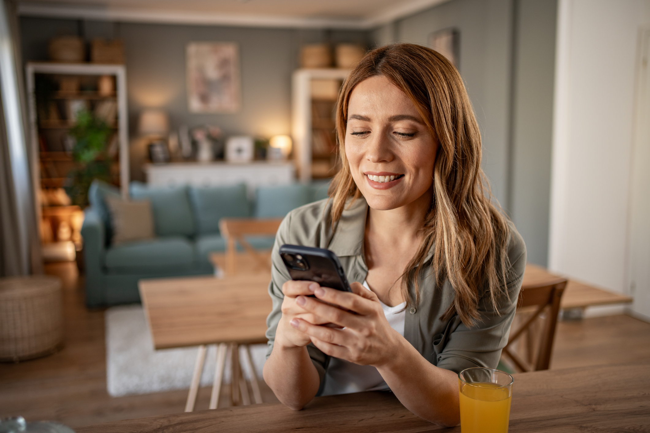 A smiling woman sitting at a table in a cosy living room, holding a smartphone while reading an SMS message, with a glass of orange juice beside her.