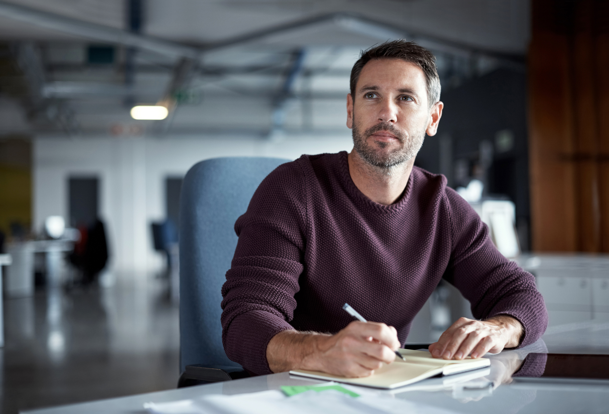 A thoughtful man sitting at a desk in an office, holding a pen and writing in a notebook, as he reflects on crafting the perfect message.