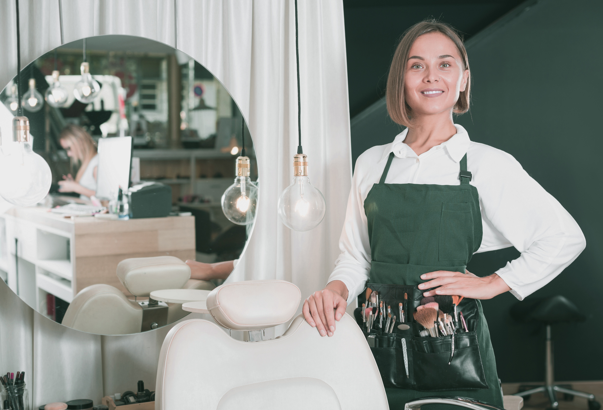 A confident female hairdresser standing in her salon, wearing an apron with styling tools, smiling and looking ready to serve clients, with a reflection in the salon mirror showing another stylist working at the desk in the background.