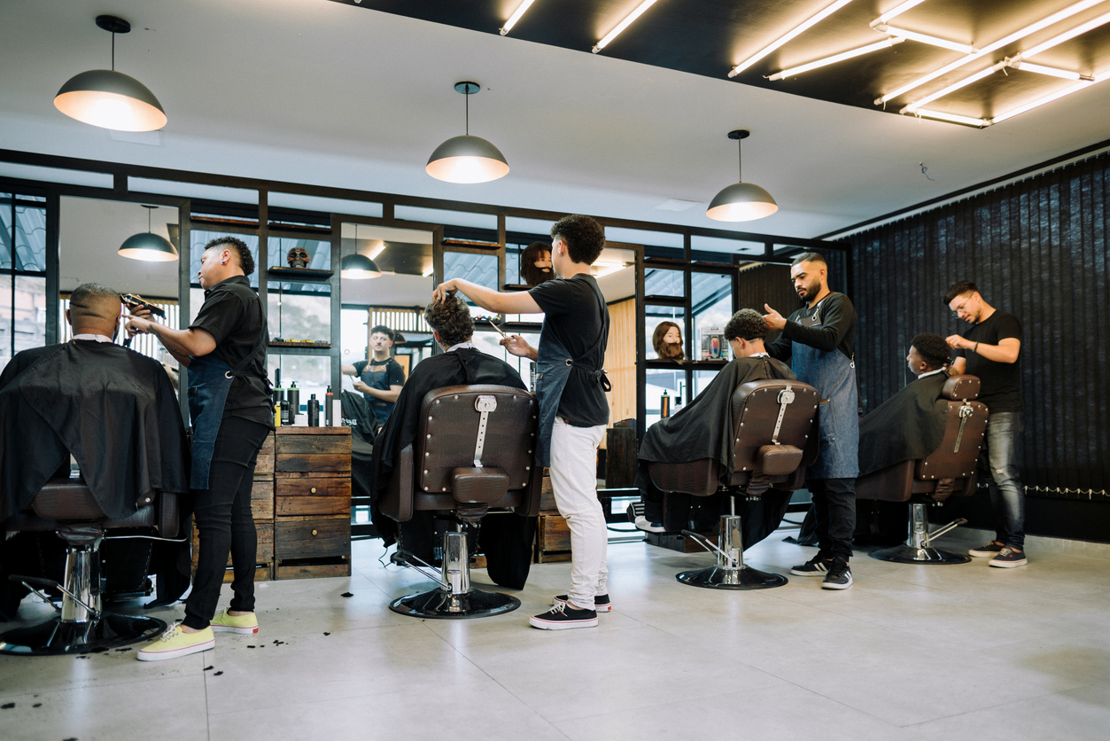 A busy barbershop with multiple stylists attending to clients. Each barber focuses on their respective client, showcasing a fully booked day at the salon—a visual representation of how effective booking systems and SMS marketing strategies can help fill every available appointment slot.