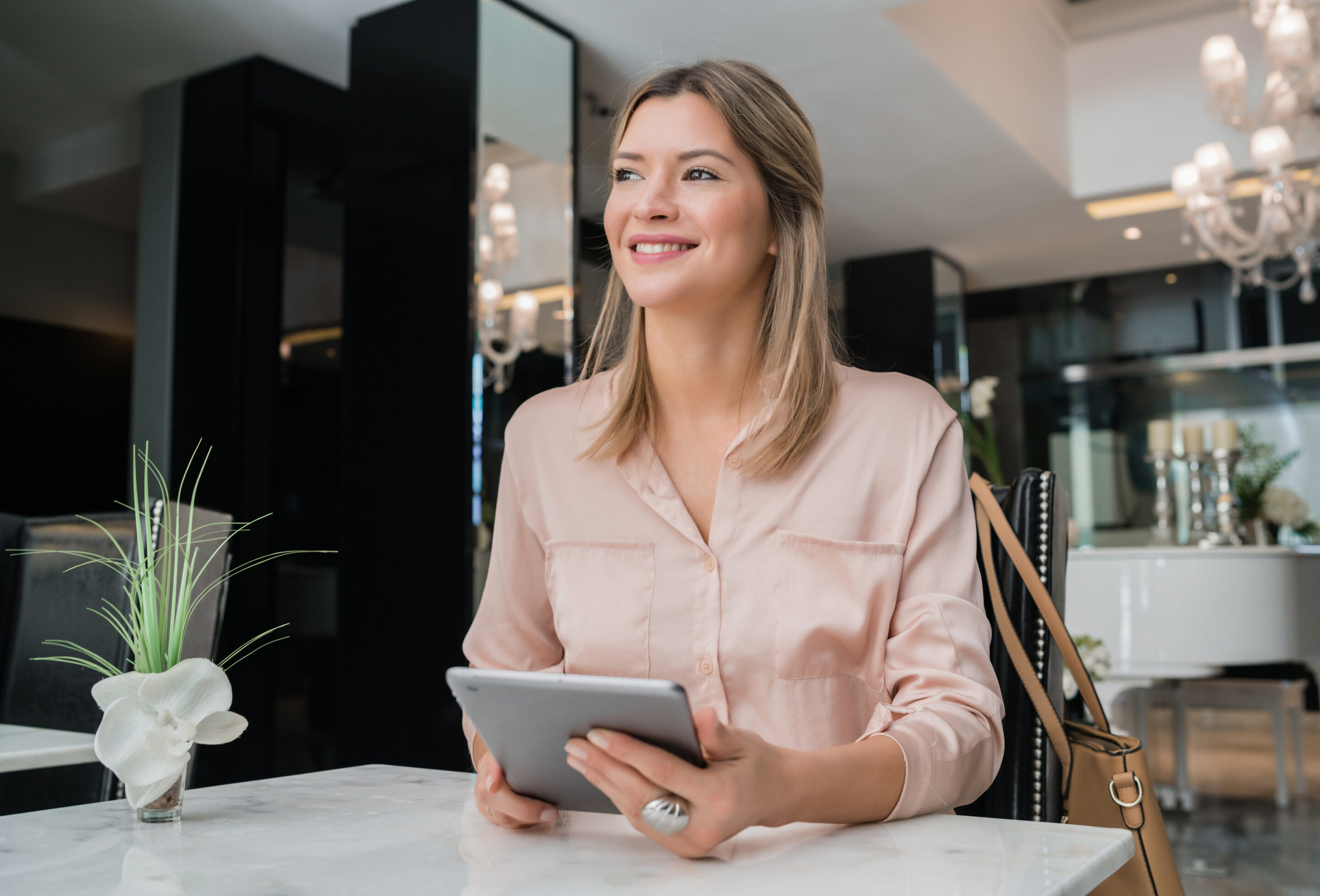 A smiling woman in a light pink blouse sits at a table in a modern salon, holding a tablet, symbolizing confidence and efficiency in managing salon operations.